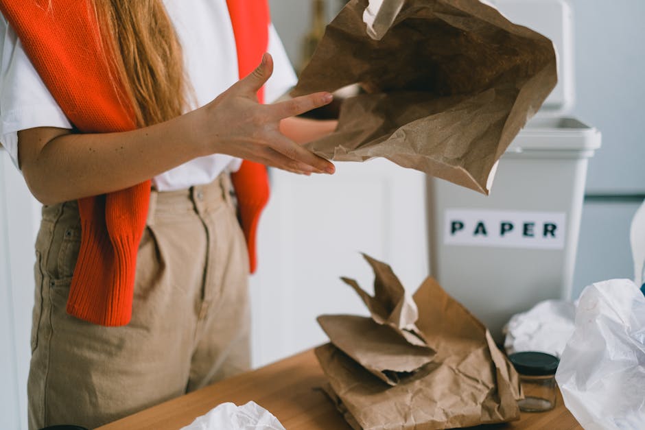 Close-up of a person's hands holding an empty brown paper bag over a cluttered table with additional crumpled paper and waste materials, positioned indoors near a white paper recycling bin labeled 'PAPER'. The individual is dressed in a white top with an orange sleeve and beige trousers, suggesting they are involved in waste sorting or collection. The background includes a white container for paper waste, indicating an environment focused on proper waste segregation and disposal. The scene emphasizes manual handling of generic waste materials typical of private rubbish collection services, aligning with concepts of on-site clearance or independent rubbish removal. The lighting appears natural and soft, highlighting the textures of the crumpled brown paper and the casual setting, which reflects a typical domestic or small business waste management scenario, suitable for guiding households on effective rubbish disposal methods in busy settings, as discussed in the page about quick rubbish collection tips.