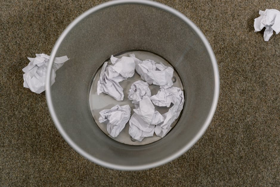 A top-down view of a cylindrical metal rubbish bin placed on a brown carpeted floor, containing several crumpled white paper balls submerged in a small amount of water. The bin's interior walls are smooth and metallic, reflecting some light. Outside the bin, additional crumpled white paper pieces are scattered on the carpet, with one crumpled paper piece positioned to the right. The environment appears to be an indoor office or workspace area, suggesting the scene relates to waste disposal or rubbish removal, potentially representing private or independent waste collection methods handled by services like Rubbish Clearance Clapham. The lighting is neutral, highlighting the textures of the paper and the metallic surface of the bin, emphasizing the contrast between the discarded waste and the surroundings, aligning with themes of rubbish clearance and alternative waste handling.