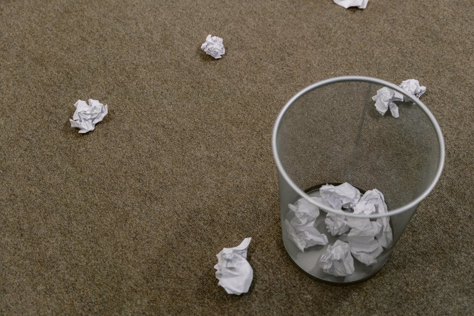 A cylindrical mesh waste bin made of transparent plastic with a metallic finish, positioned on a brown carpeted floor. The bin contains crumpled white paper and is surrounded by several additional crumpled pieces of paper scattered on the floor, indicating a cluttered workspace or office environment. The scene is lit with soft, natural light, highlighting the textures of the crumpled paper and the carpet surface. This setup suggests a context of manual or independent rubbish collection, aligning with services such as those offered by Rubbish Clearance Clapham for on-site or private waste disposal in a professional rubbish removal setting.