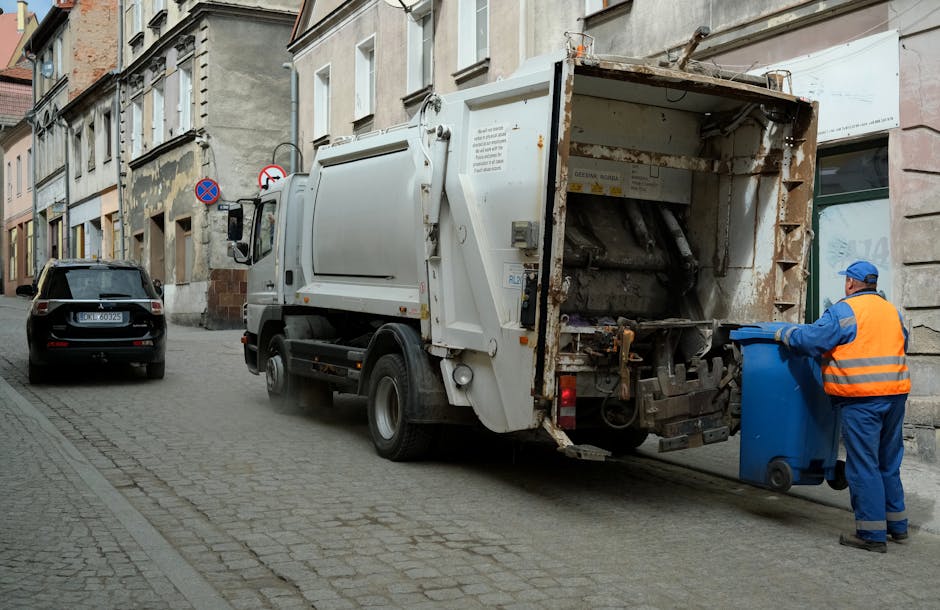 A sanitation worker dressed in blue trousers, a high-visibility orange vest, and a blue cap is standing on a cobblestone street next to a large white rubbish collection truck with an open rear loading area that shows rust and wear. The worker is holding a blue wheeled bin, positioning it to be emptied into the truck. The truck is parked parallel to the pavement, which is lined with multi-storey residential buildings featuring stone and brick facades, with some windows and architectural details visible. In the background, there is a black car parked further along the street, and traffic signs indicating parking restrictions. Natural daylight illuminates the scene, providing clear visibility of the environment, the materials and textures of the rubbish truck, the metal and rusted parts, as well as the brick and stone surfaces of the surrounding buildings. The scene reflects an aspect of private waste collection and on-site rubbish removal services, typical of independent or commercial waste handling operations in urban areas. Rubbish Clearance Clapham may handle similar collections for residents requiring alternative rubbish disposal options.