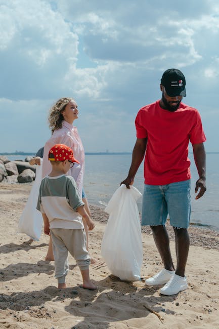 A man with dark skin, wearing a red T-shirt, light blue denim shorts, white sneakers, and a navy baseball cap, is standing on a sandy beach holding a white trash bag, preparing to dispose of rubbish. Behind him, a woman with curly blonde hair, dressed in a pink jacket, and a young boy wearing a red cap with yellow patterns, a light gray T-shirt, and beige trousers, are also on the beach, with the woman looking towards the water and the boy facing away from the camera. The scene is set by a body of water with rocky shoreline elements visible in the background. The cloudy sky creates a diffuse lighting atmosphere, and the environment appears calm and natural. This scene reflects a community or family effort to clean up or perform a private rubbish collection service, illustrating the importance of waste disposal at outdoor locations, relevant to independent rubbish removal activities often promoted by local waste management services like Rubbish Clearance Clapham.