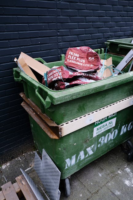 A green wheeled waste container filled with bags of roofing materials, including red and black flexible pipe insulation, stacked on top of a second green container against a dark brick wall. The second container has a white protective edging strip and some printed labels. The scene is outdoors on a paved surface, with part of a wooden pallet and a metal grate visible at the bottom left corner. The area appears to be a private property or service yard, with the containers used for rubbish or materials disposal, consistent with private waste collection or onsite clearance services.