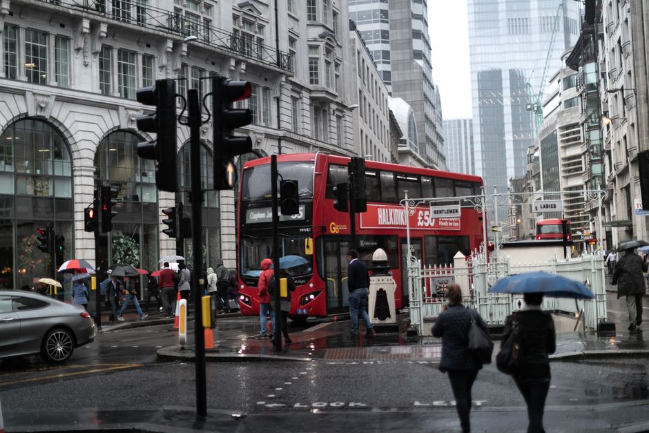 A busy urban street scene on a rainy day featuring a red double-decker bus stopping at a designated area, surrounded by pedestrians holding umbrellas walking across a wet crosswalk. The background includes tall modern office buildings with glass facades and older stone-fronted structures, creating a contrast between contemporary and historic architecture. Traffic lights and street signs are visible, with one advertising a show for 'Hamilton' on the side of the bus. Vehicles are seen on the wet roads, and some pedestrians are waiting on the sidewalk near a black cast-iron fence, indicating a typical city environment. The overall atmosphere is damp, with reflections of the umbrellas and vehicles visible on the slick road surface. This scene exemplifies an active city center with public transportation and pedestrian activity, relevant to the context of waste management and rubbish removal services operating within urban environments.