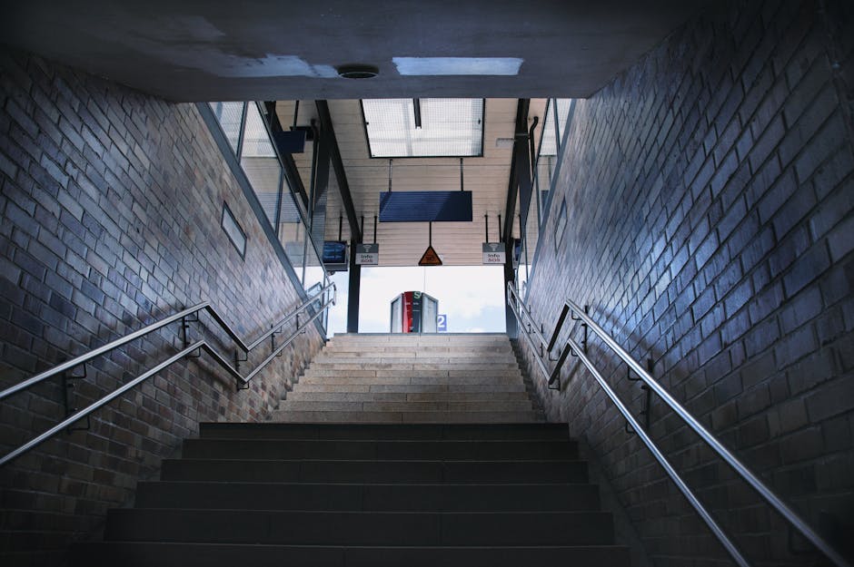 A set of concrete stairs ascending from an underground or lower-level exit of a station, framed by brick walls on either side with a textured surface and slight variation in brownish-gray tones. The stairs are fitted with metal handrails on both sides, extending from the bottom to the top, and are illuminated by natural light coming from the open exit at the top, where a glimpse of the outside can be seen. Above the stairs, a ceiling with a few small, circular light fixtures is visible, along with hanging signs indicating directions and station information. The surrounding environment suggests an urban railway station or similar transportation hub, with the scene emphasizing the structural elements and functional design typical of access points for public transportation, which are sometimes involved in on-site waste or rubbish removal activities requiring temporary storage or clearance, as managed by companies like Rubbish Clearance Clapham.