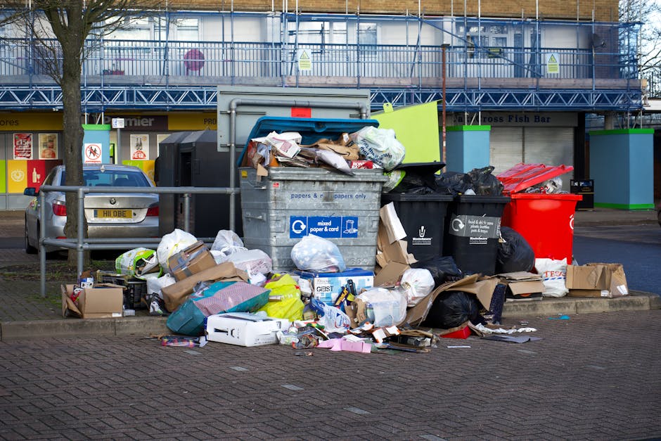 A large collection of mixed waste and rubbish bags, including black plastic trash bags, brown cardboard boxes, and various paper and packaging materials, are piled on a paved area beside a metal rubbish bin with a blue lid in an urban setting. Some rubbish is overflowing from the open bin, with items spilling onto the ground. The scene is situated near a parking space with a grey car parked on the left, and a metal barrier fence separates the rubbish area from the surrounding pavement. In the background, there is a building with a yellow shopfront, signage, and a closed shutter, with scaffolding and safety warning signs visible on the upper levels. The environment suggests a city street or shopping area, where private waste collection or disposal services might be used to manage overflows beyond scheduled municipal rubbish collection for local waste removal services. The lighting appears natural, with overcast skies casting diffuse light over the scene, highlighting the textures of the various waste materials and surfaces.